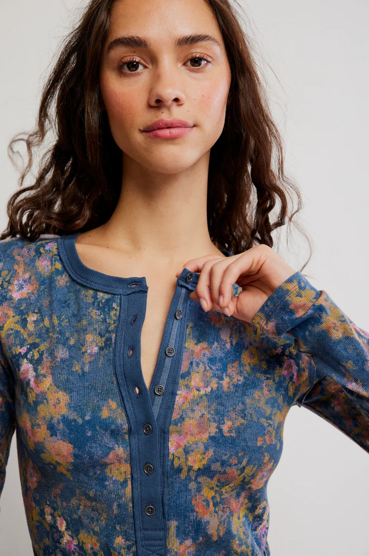 A close-up of a woman wearing a long-sleeve button-front top with a blue base and a multicolor floral print in pink, yellow, and green, against a light gray background.