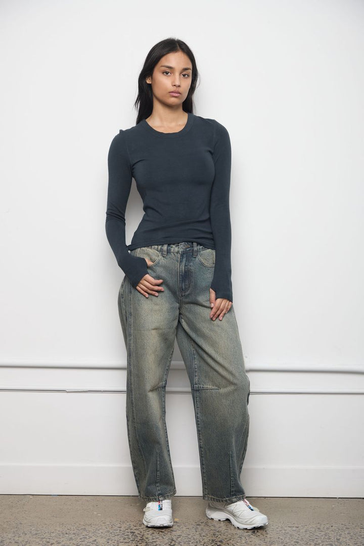 A model stands against a white wall wearing a dark charcoal long-sleeve top, wide-leg faded gray jeans, and white sneakers.