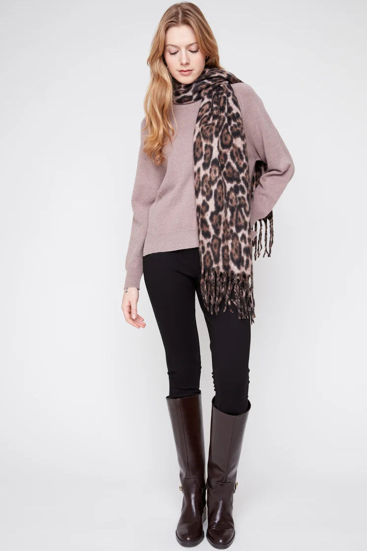 A woman modeling a leopard print scarf with fringe, styled with a mauve sweater, black pants, and dark brown boots against a light gray studio background.