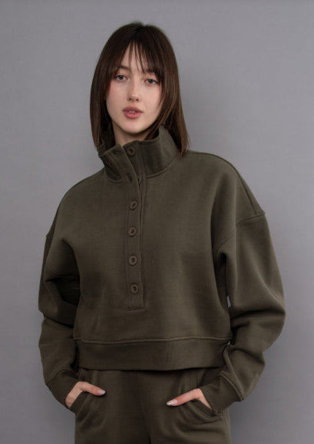 A woman modeling an olive green cropped quarter-zip sweatshirt and matching sweatpants against a solid gray studio background.