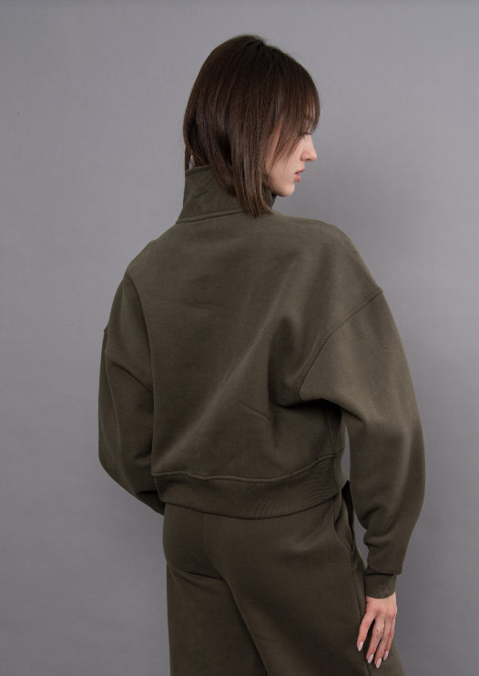A model shown from the back wearing an oversized olive green mock-neck sweatshirt and matching sweatpants against a solid gray studio background.