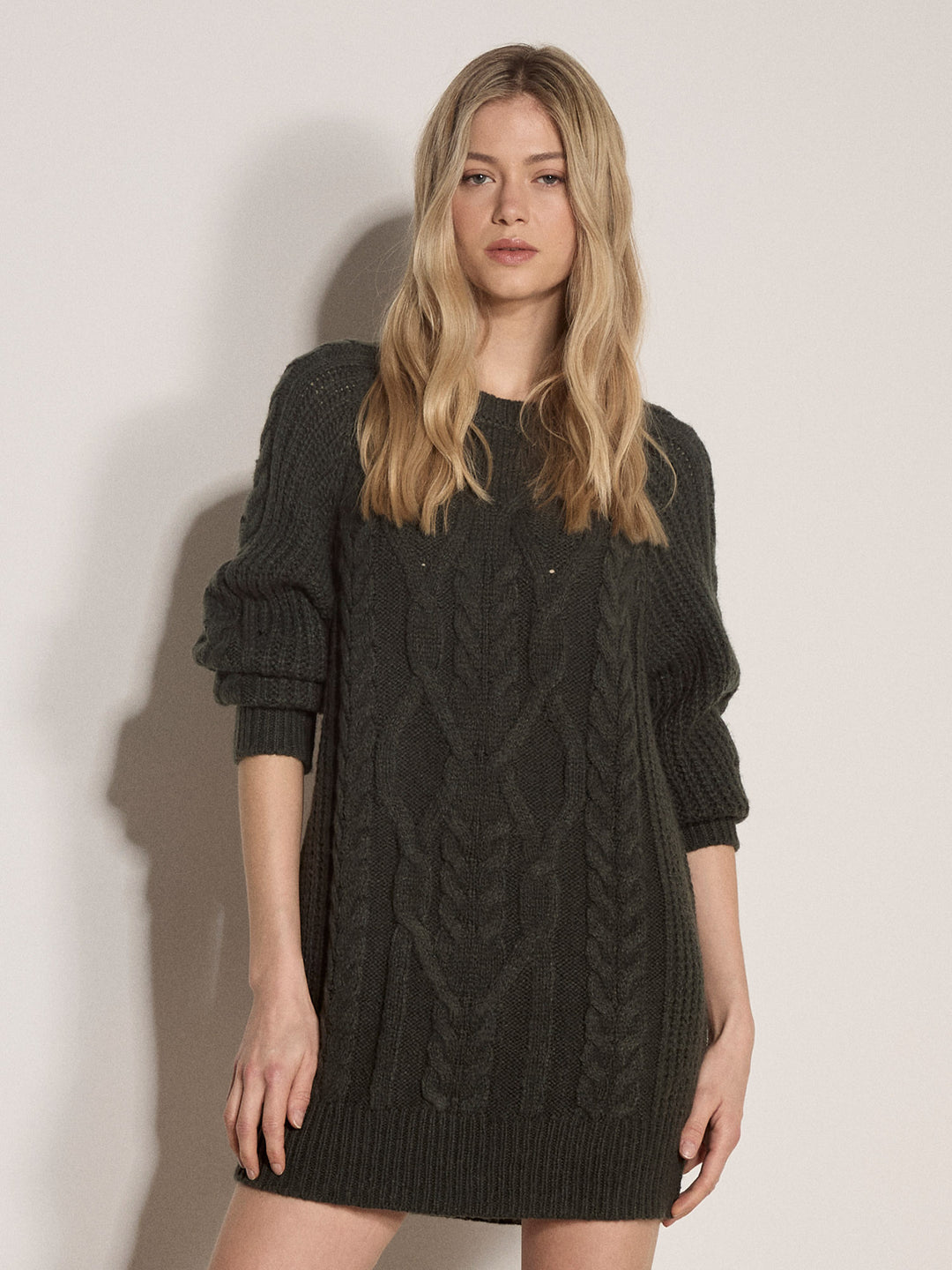 A blonde woman wearing a dark charcoal cable-knit sweater dress, standing against a plain off-white wall.