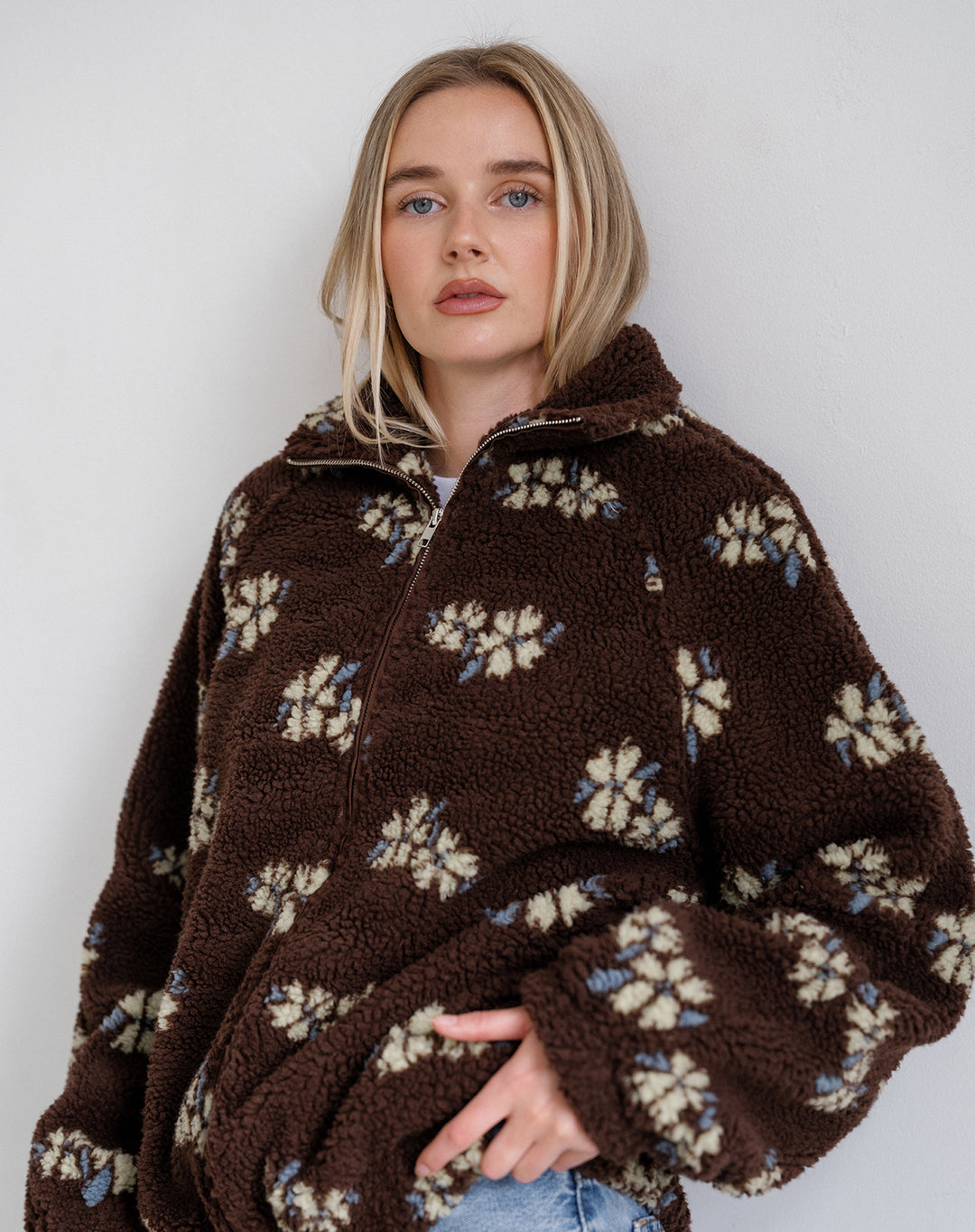 A woman wearing a brown sherpa fleece quarter-zip jacket with a cream and light blue floral pattern, posed against a plain light gray background.
