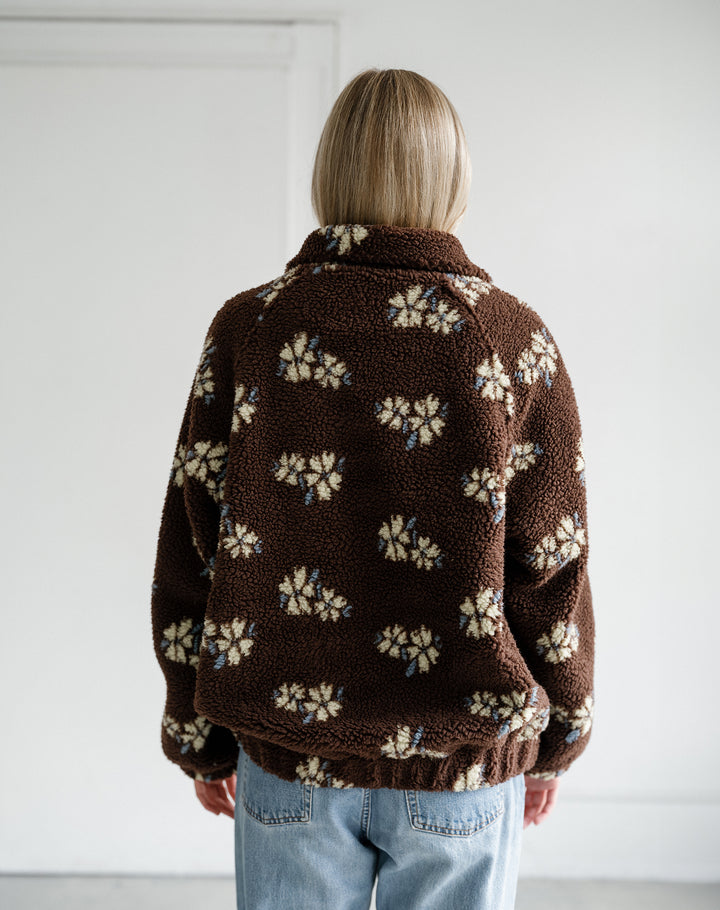 A person viewed from behind wearing a brown sherpa fleece jacket with a cream and light blue floral pattern, paired with light blue jeans, against a plain white wall.