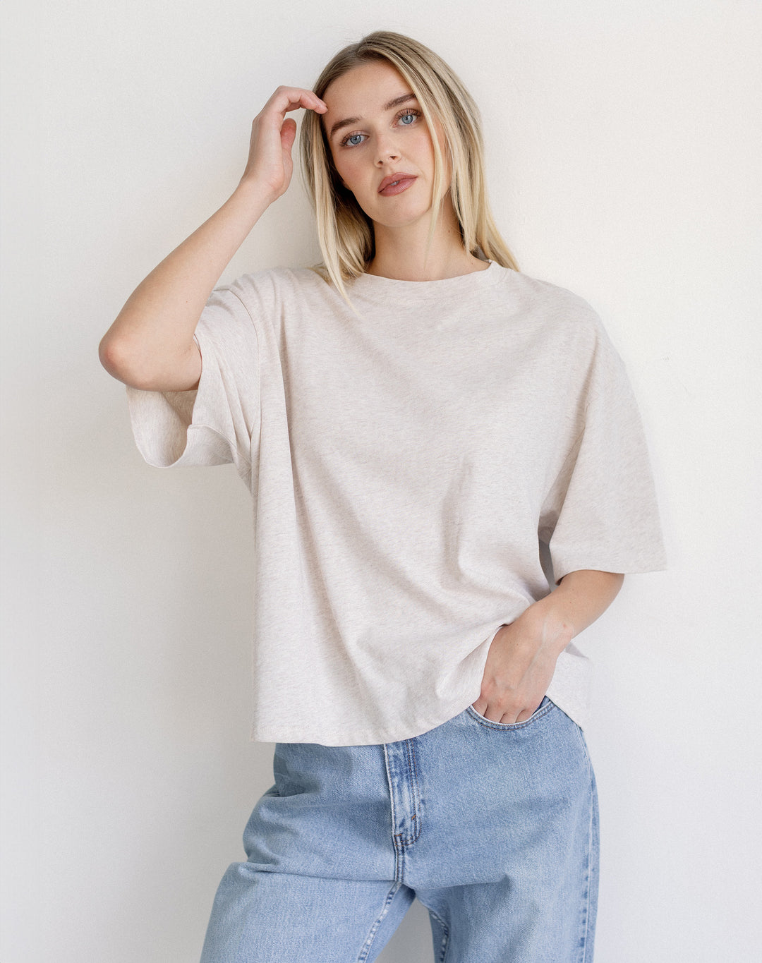 A woman wearing an oversized short-sleeve t-shirt in a light oatmeal beige color, paired with light blue jeans, posing against a plain white wall.