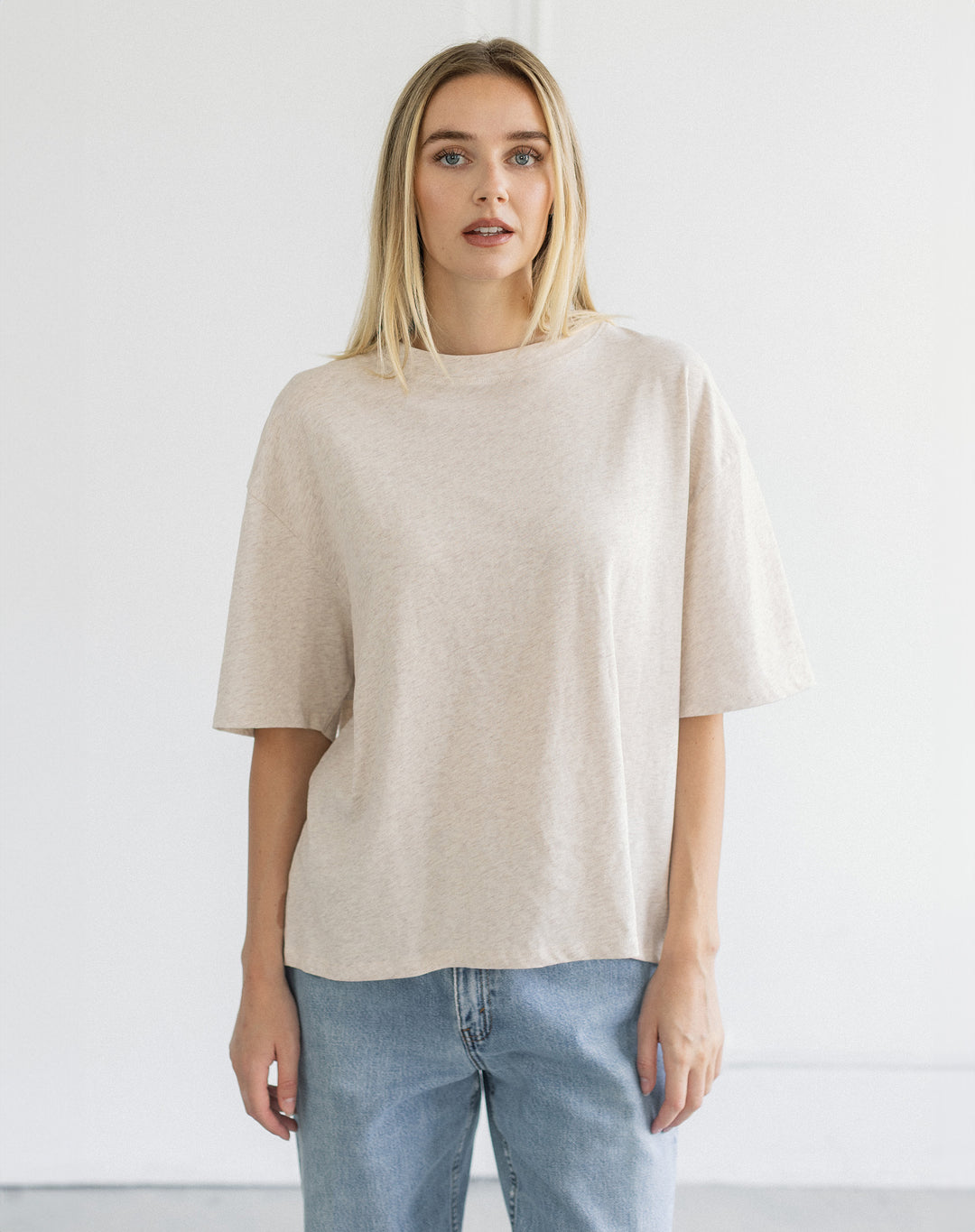 A blonde woman wearing a loose, short-sleeve beige knit top and light blue jeans, standing against a plain white studio background.