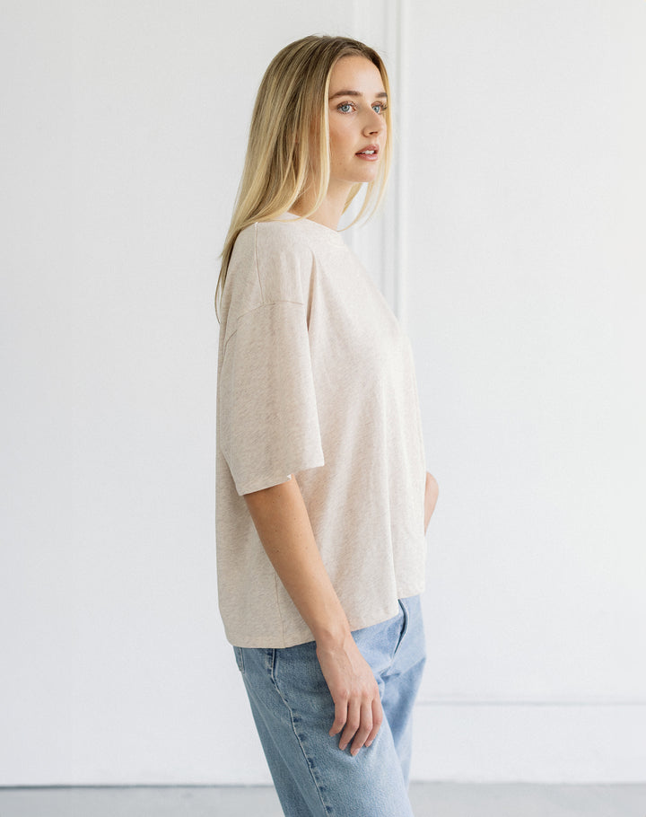 A woman with blonde hair is shown in profile wearing a light beige short-sleeve knit top and light blue jeans against a bright white studio background.