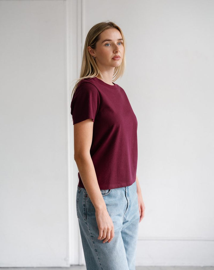 A woman wearing a short-sleeve crewneck t-shirt in burgundy and light wash blue jeans, standing in a minimalist white room.
