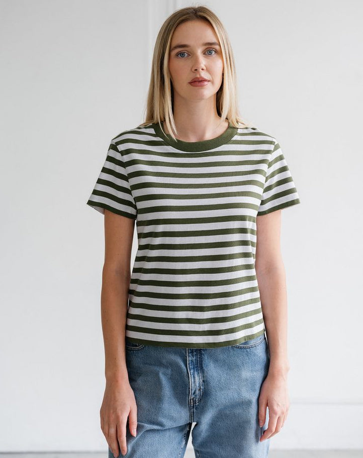 A woman modeling a short-sleeve crewneck t-shirt with horizontal olive green and off-white stripes, paired with light-wash blue jeans, set against a plain white wall.