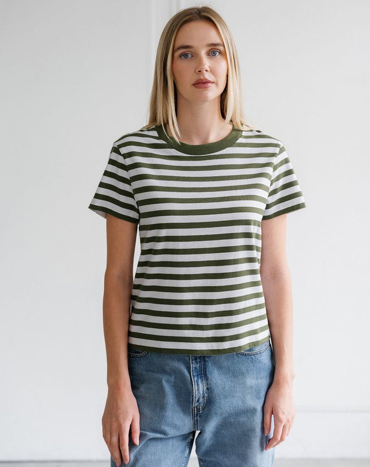 A woman modeling a short-sleeve crewneck t-shirt with horizontal olive green and off-white stripes, paired with light-wash blue jeans, set against a plain white wall.