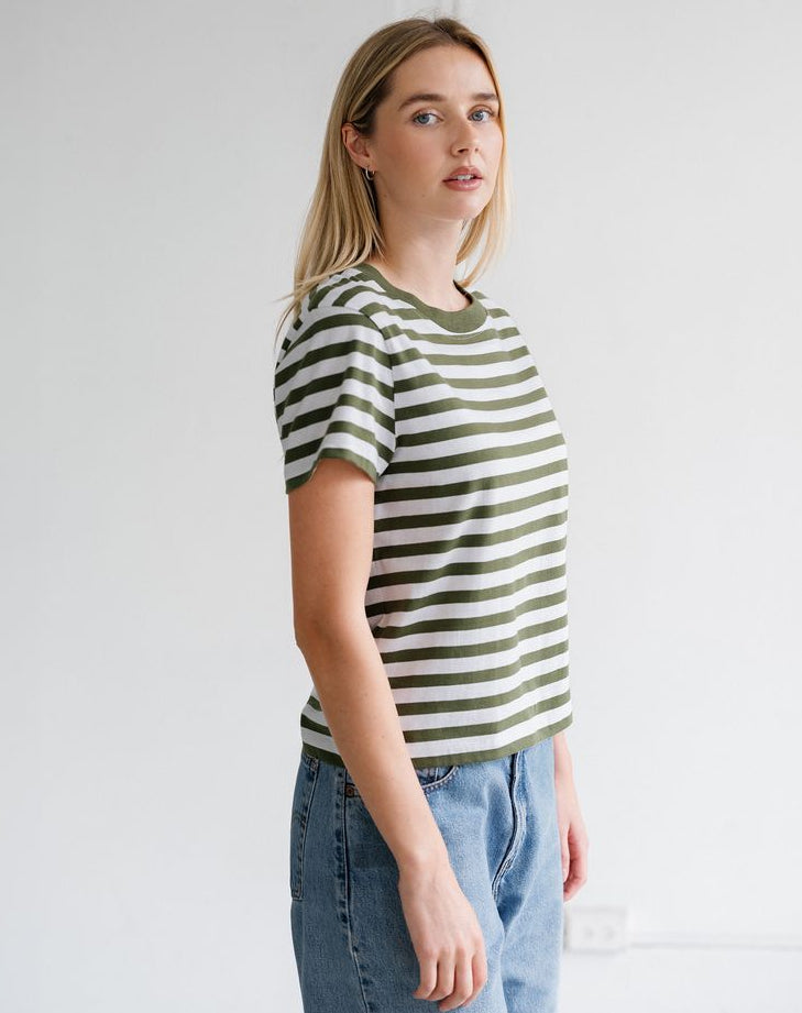 A woman modeling a short-sleeve crewneck t-shirt with horizontal olive green and off-white stripes, paired with light wash blue jeans, standing against a plain white wall.