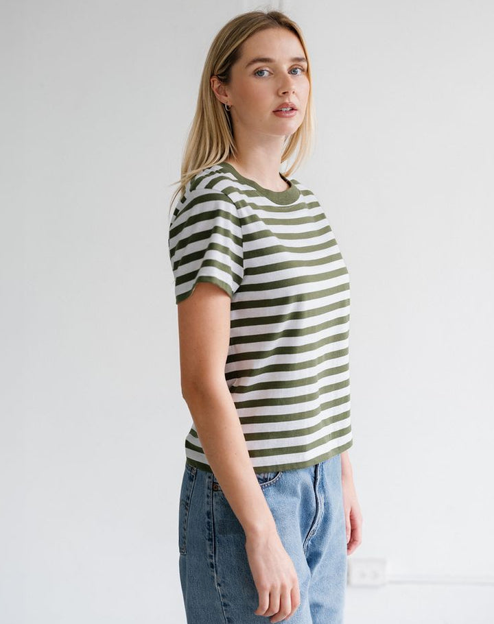 A woman modeling a short-sleeve crewneck t-shirt with horizontal olive green and off-white stripes, paired with light wash blue jeans, standing against a plain white wall.