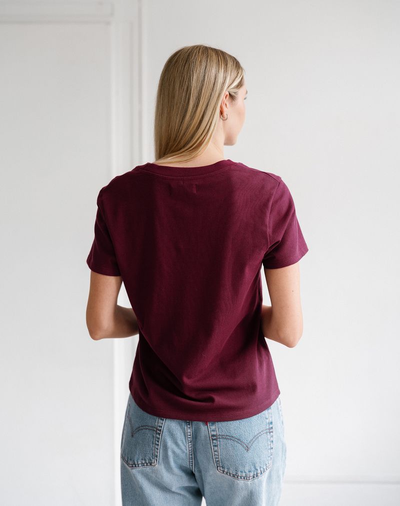A woman viewed from behind wearing a short-sleeve crewneck t-shirt in deep burgundy and light wash blue jeans, standing against a plain white wall.