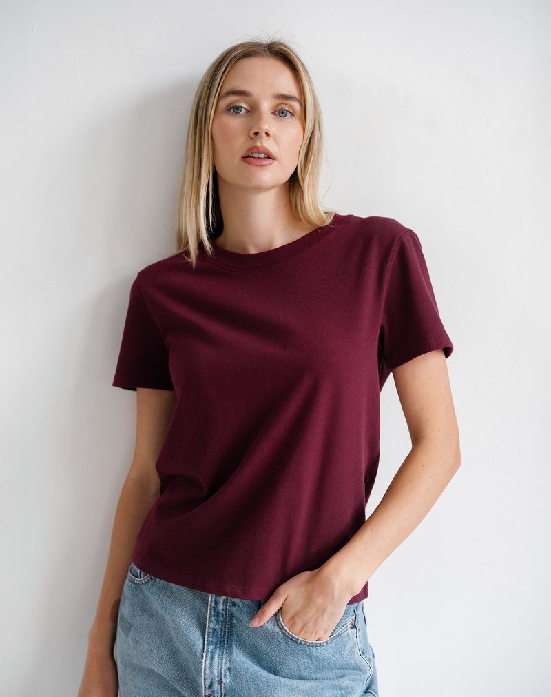 A woman wearing a short-sleeve crewneck t-shirt in deep burgundy, paired with light wash blue jeans, standing against a plain white wall.