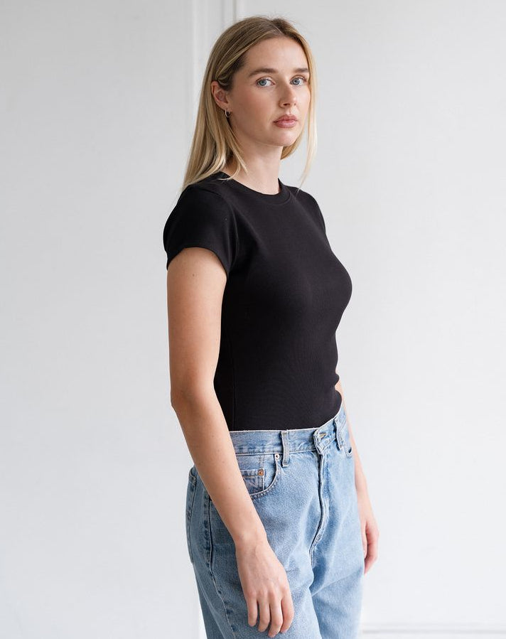 A woman wearing a fitted black short-sleeve crewneck top and light wash blue jeans, standing against a plain white wall.