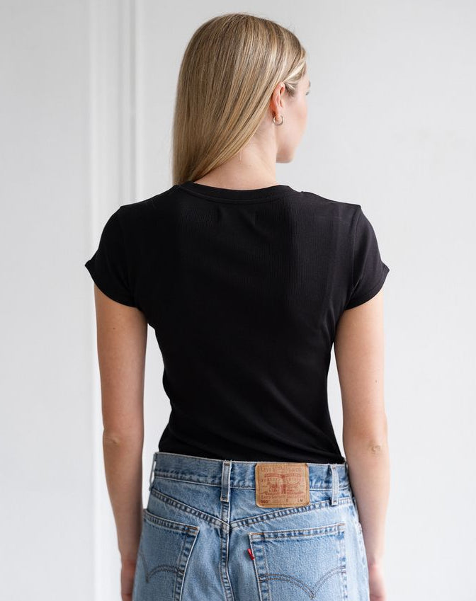 A woman viewed from behind wearing a fitted black short-sleeve t-shirt and light blue jeans with a visible leather patch on the back waistband.