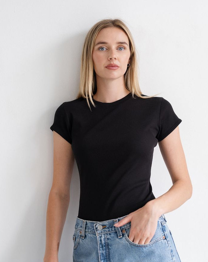 A blonde woman wearing a fitted black short-sleeve crewneck t-shirt and light-wash blue denim jeans, standing against a plain white wall.