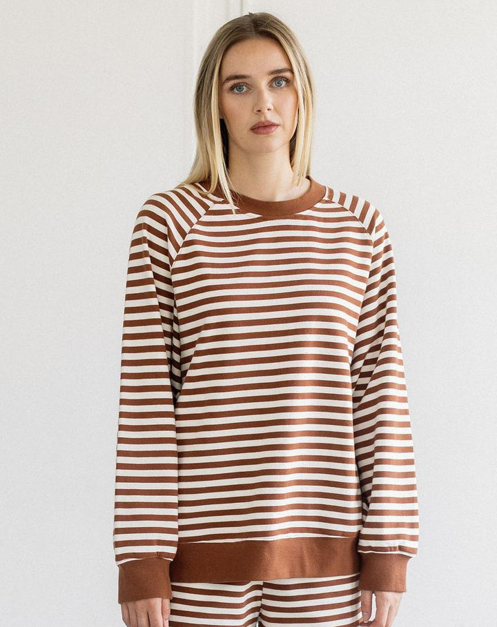 A woman modeling a matching two-piece lounge set consisting of a long-sleeve crewneck sweatshirt and shorts, both featuring horizontal brown and off-white stripes with solid brown ribbed trim.