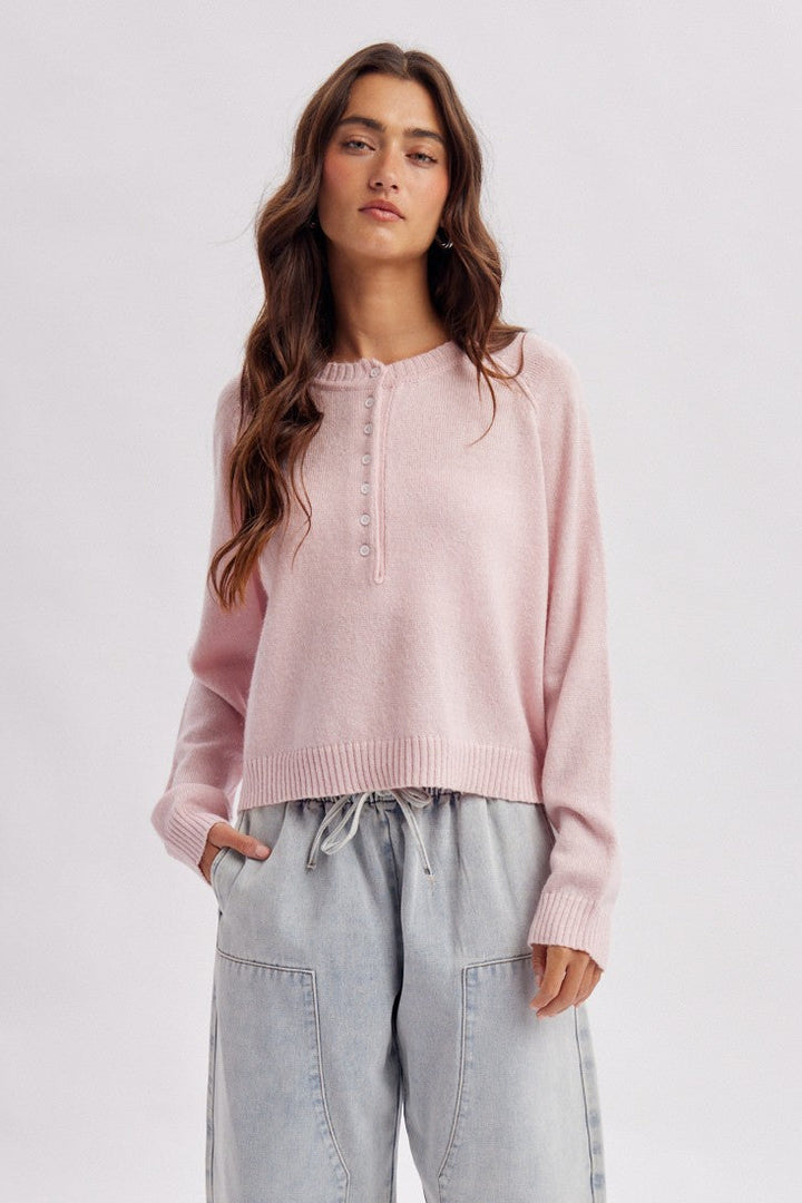 A woman modeling a light pink long-sleeve knit sweater with a buttoned henley-style neckline, paired with wide-leg light gray drawstring jeans and white flat shoes, against a plain light gray studio background.