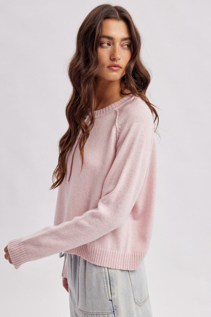 A woman modeling a light pink knit crewneck sweater paired with light gray jeans, photographed against a solid light gray studio background.