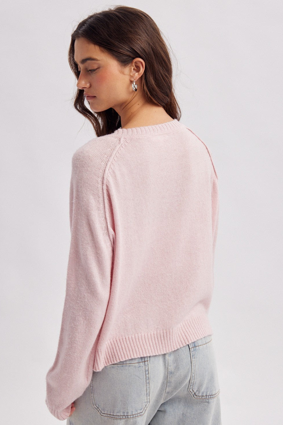 A woman wearing a light pink knit sweater and light blue jeans, shown from a side-back angle against a plain light gray studio background.