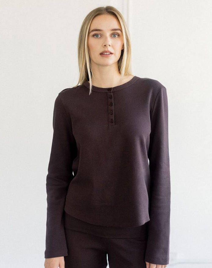 A woman modeling a dark plum long-sleeve henley top with matching dark pants against a plain white studio background.