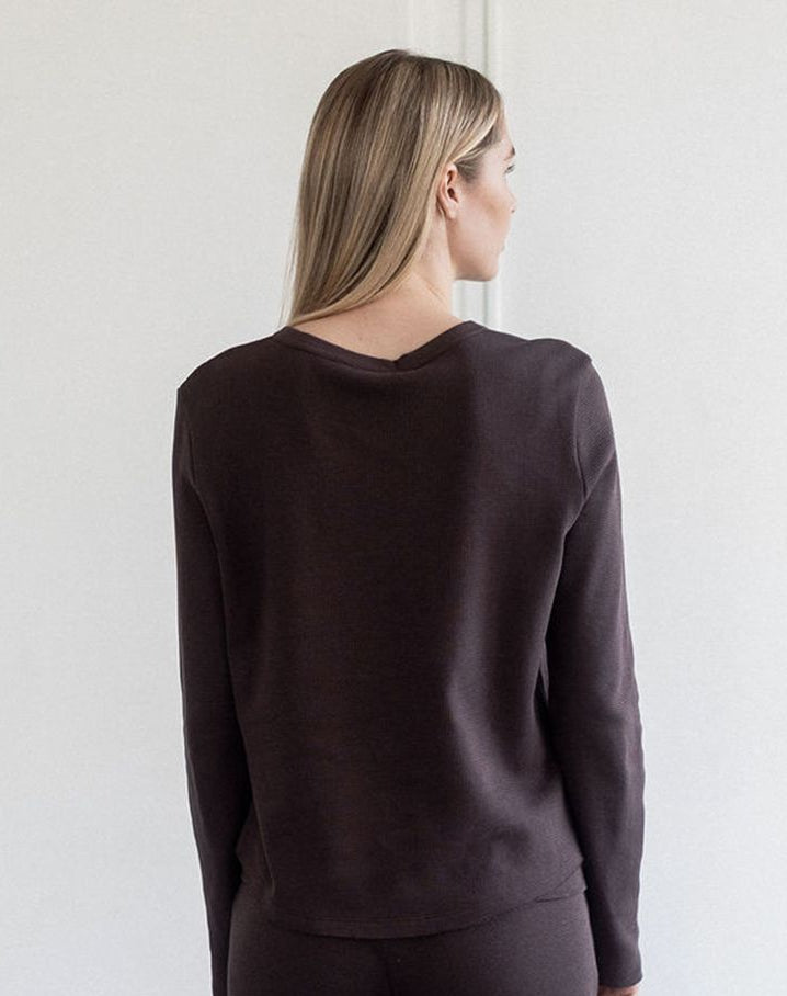 A woman shown from the back wearing a dark brown long-sleeve knit top and matching dark pants against a plain light gray wall.