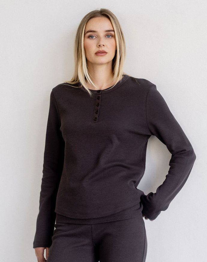 A woman modeling a dark charcoal long-sleeve henley top with a buttoned neckline, paired with matching dark leggings, against a plain light gray background.
