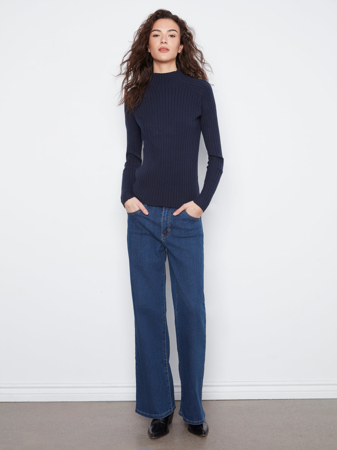 A woman modeling a dark navy ribbed mock-neck long-sleeve sweater paired with blue wide-leg jeans and black shoes, standing against a white wall.
