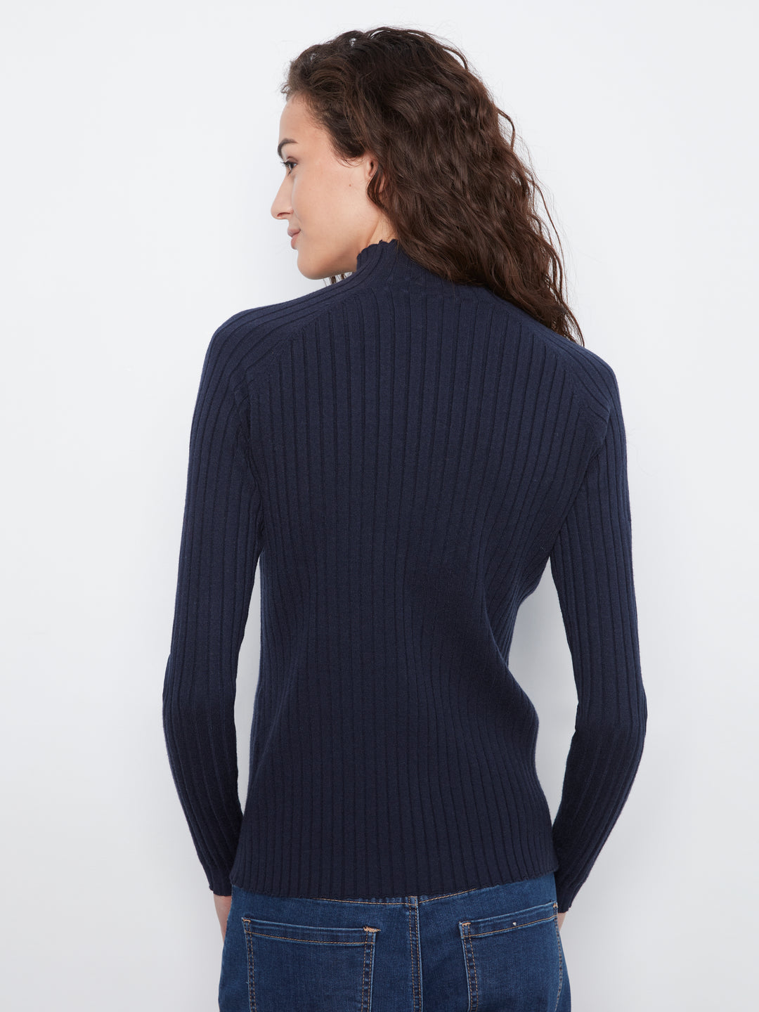 A woman with long dark curly hair is shown from the back wearing a navy blue ribbed knit mock neck sweater and blue jeans against a plain white background.