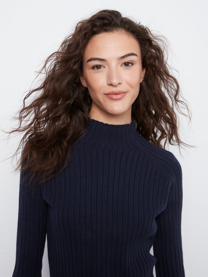 A woman with long, wavy brown hair wearing a dark navy ribbed mock-neck long-sleeve sweater, photographed against a clean white studio background.
