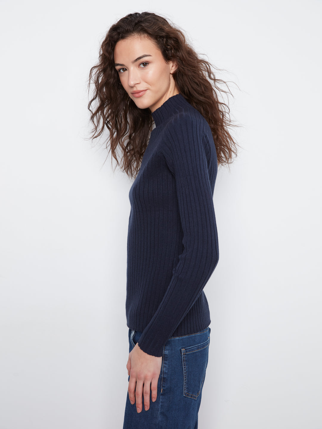 A woman with long, wavy brown hair is shown in profile wearing a dark navy ribbed knit mock-neck sweater and blue jeans against a plain white studio background.
