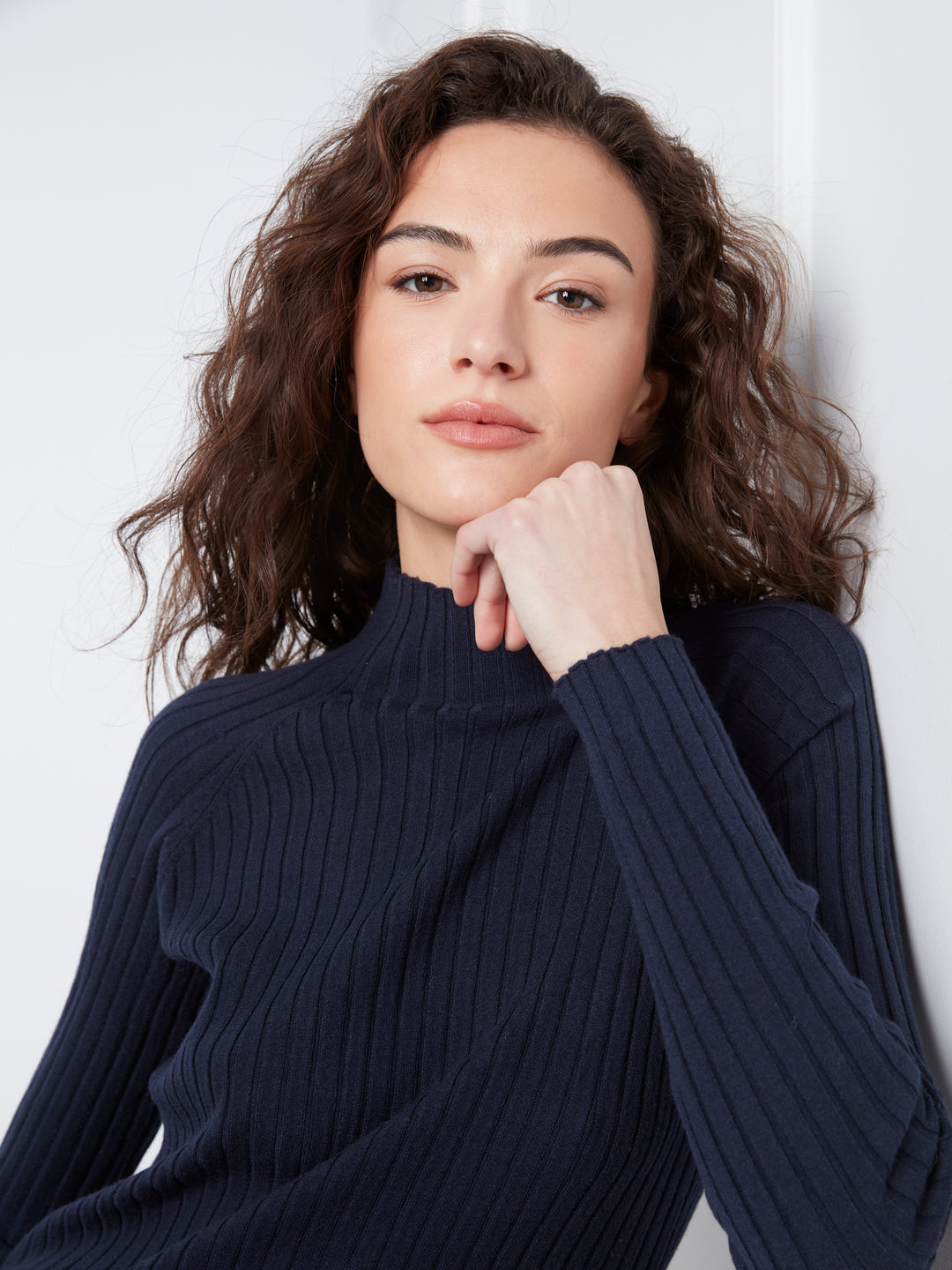 A woman with wavy brown hair wearing a dark navy ribbed mock neck sweater, posing with her hand resting on her chin against a light gray background.