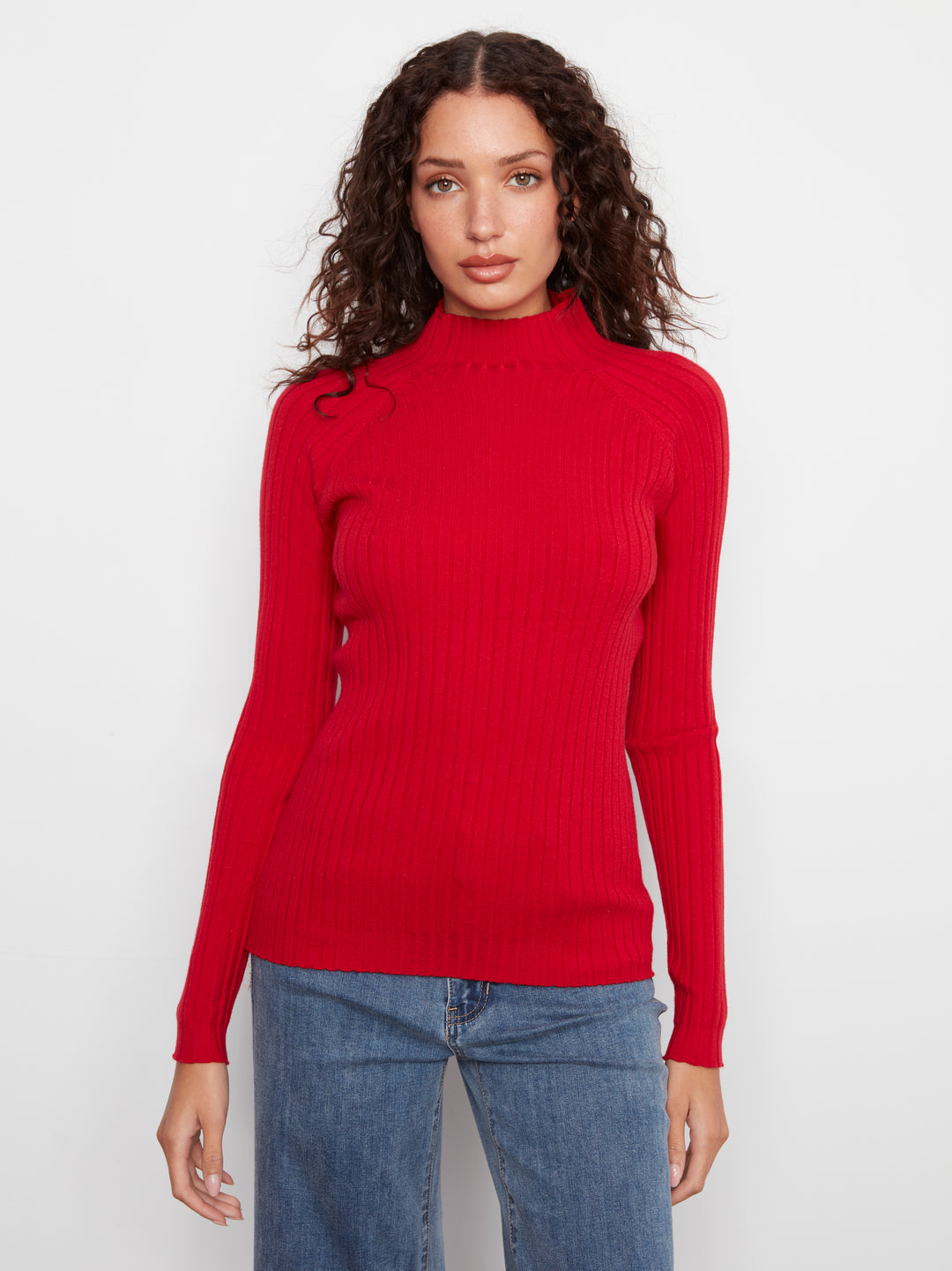 A woman with curly brown hair wearing a bright red ribbed mock neck sweater and blue jeans, standing against a plain white studio background.