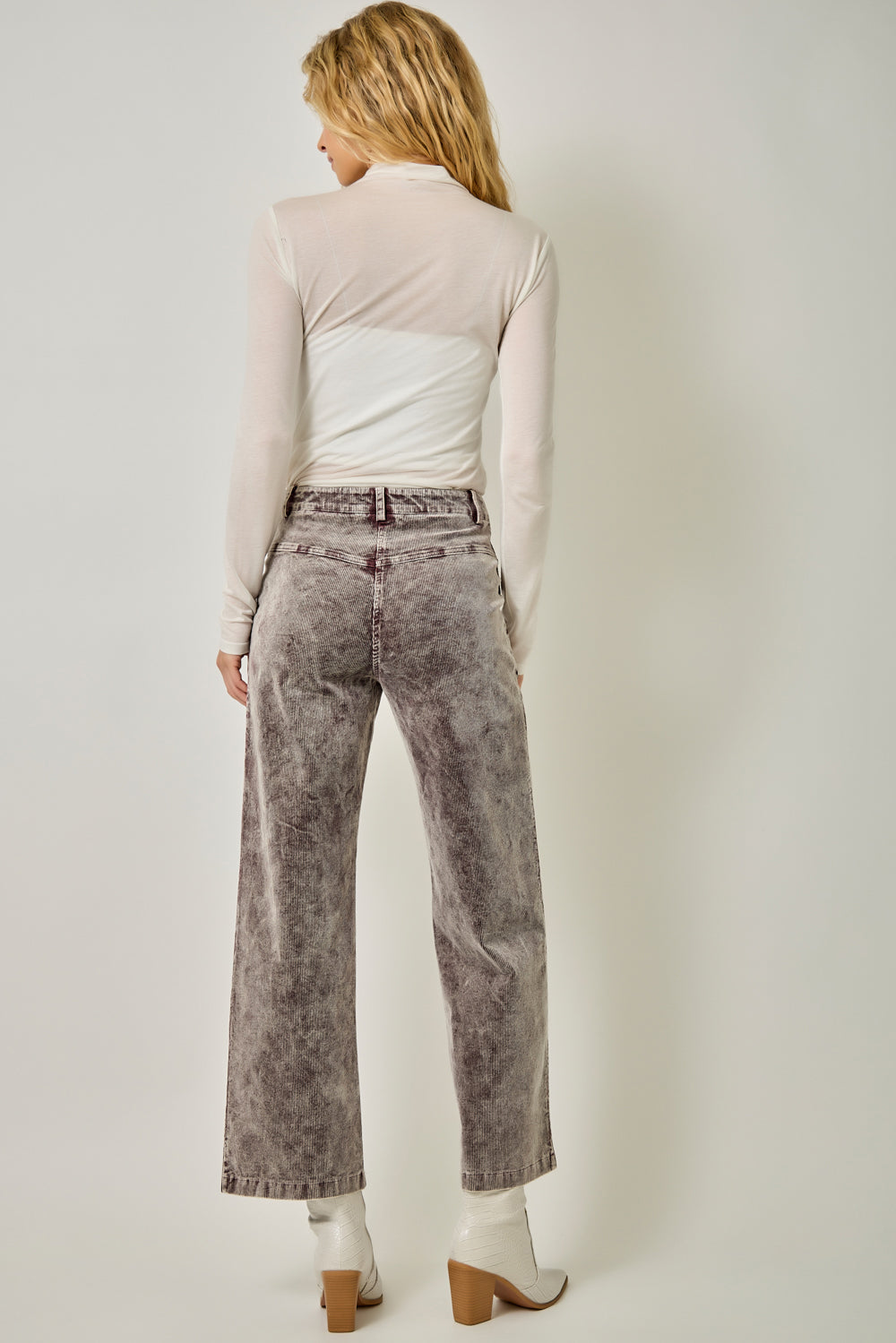 A woman shown from the back wearing a sheer long-sleeve top, gray acid-wash wide-leg jeans, and white heeled boots against a plain light gray studio background.