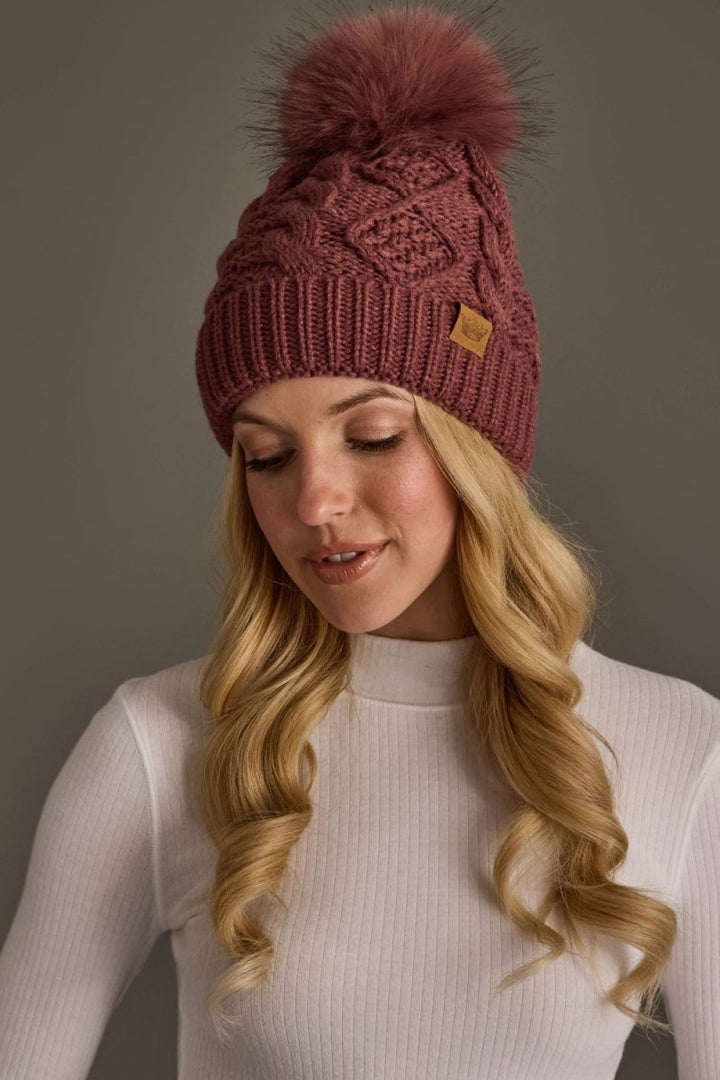 A woman wearing a dusty rose cable-knit beanie with a large matching faux fur pom-pom and a small tan brand tag, paired with a white ribbed mock-neck top against a neutral gray background.