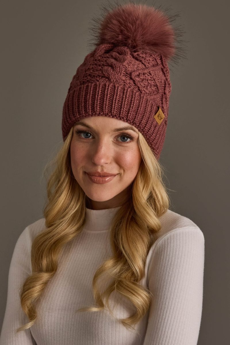 A woman with long blonde hair wearing a dusty rose cable-knit beanie with a matching faux-fur pom-pom and a small tan diamond logo, posed against a solid dark gray background.