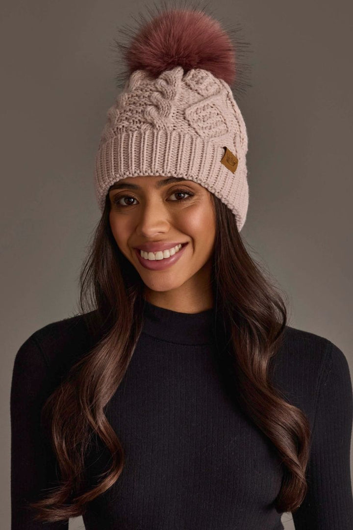 A smiling woman wearing a light pink cable-knit beanie with a large dusty rose pom-pom and a small brown leather logo tab, paired with a dark navy mock-neck top against a neutral gray studio background.