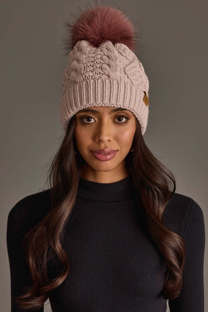 A woman wearing a cable-knit beanie with a large dusty rose faux fur pom-pom and a small gold logo on the side, paired with a black ribbed mock-neck top against a neutral gray background.