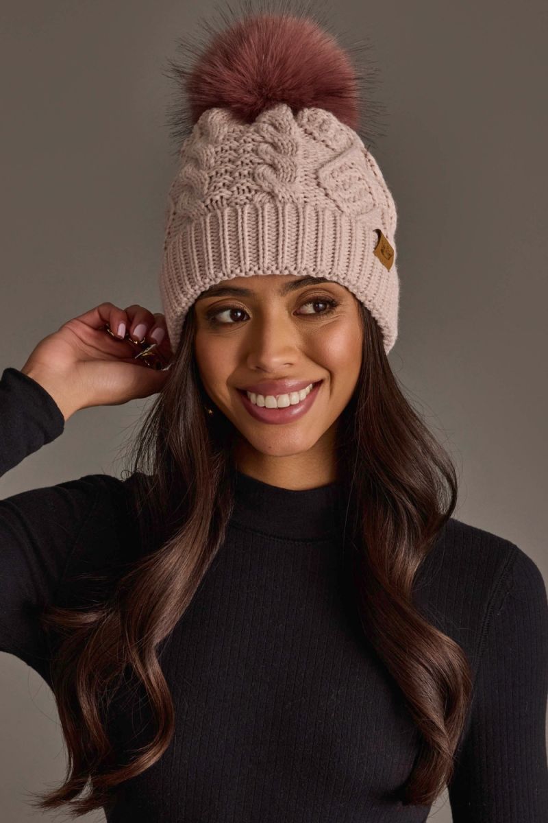A smiling woman wearing a beige cable-knit beanie with a dusty rose pom-pom and a small brown logo patch, paired with a black turtleneck sweater against a solid gray studio background.