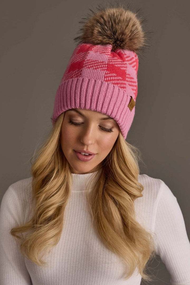 A woman wearing a pink knit beanie with a large brown faux-fur pom-pom and a small brown leather brand tab, photographed against a plain taupe background.
