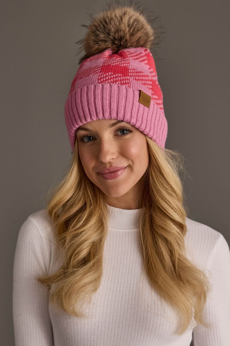 A woman wearing a pink knit beanie with a brown faux-fur pom-pom and a small tan brand patch on the side, photographed against a plain gray studio background.