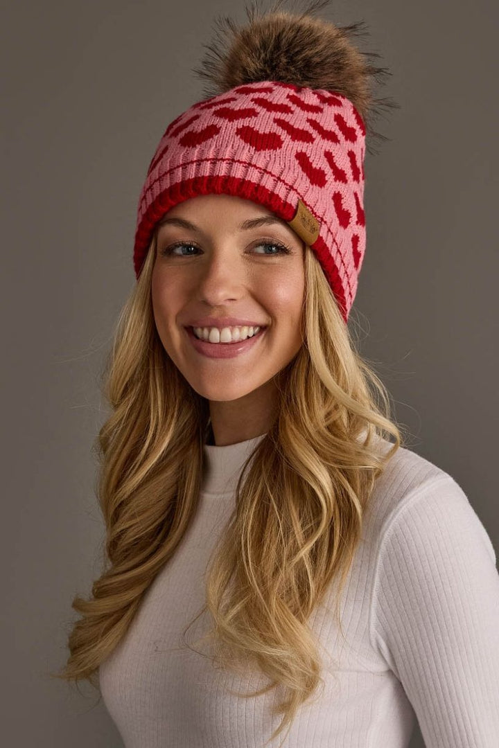 A smiling blonde woman wearing a pink and red knit beanie with a brown faux-fur pom-pom, photographed against a plain gray studio background.