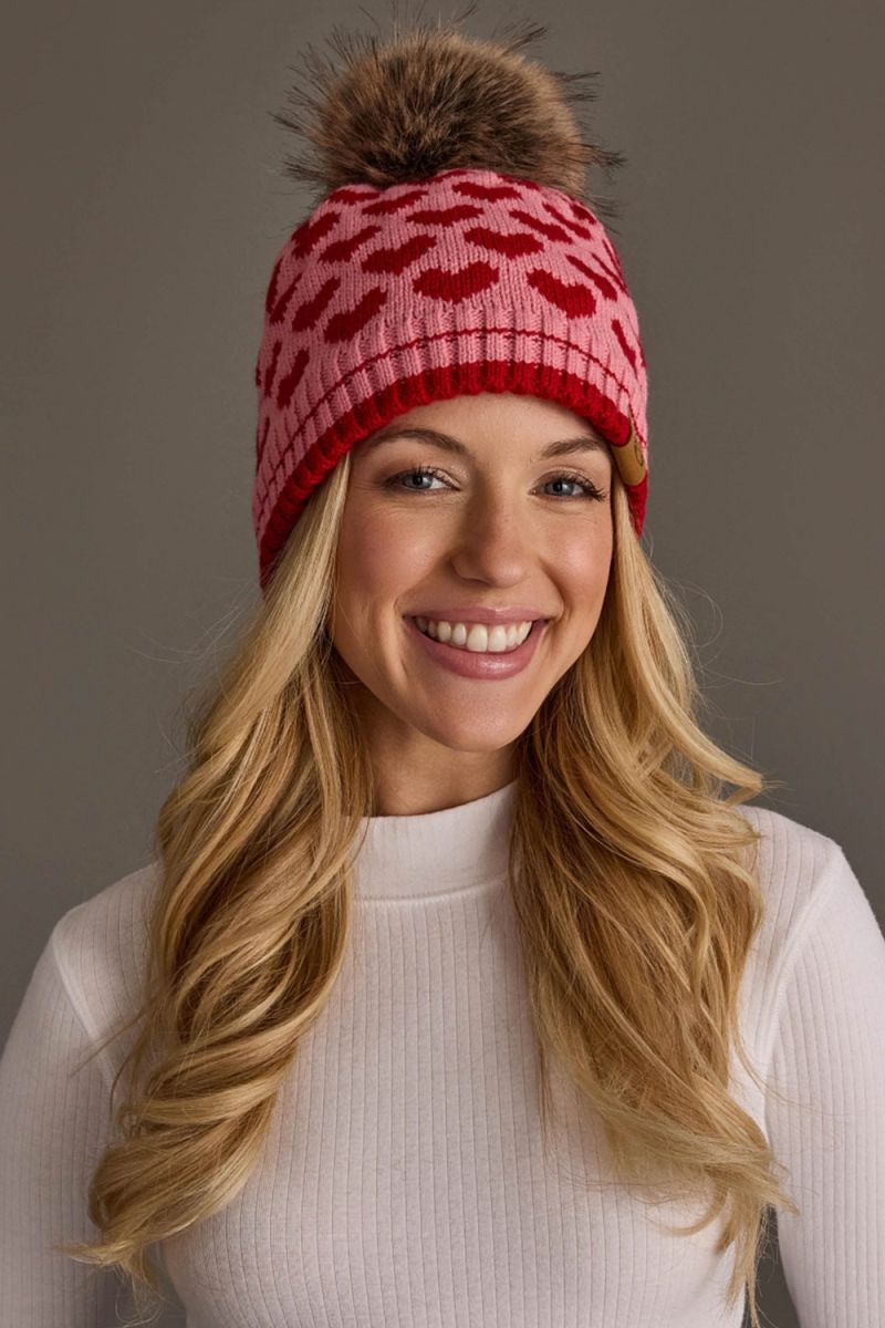A smiling blonde woman wearing a pink knit beanie with a red heart pattern and a brown faux-fur pom-pom, against a plain gray studio background.