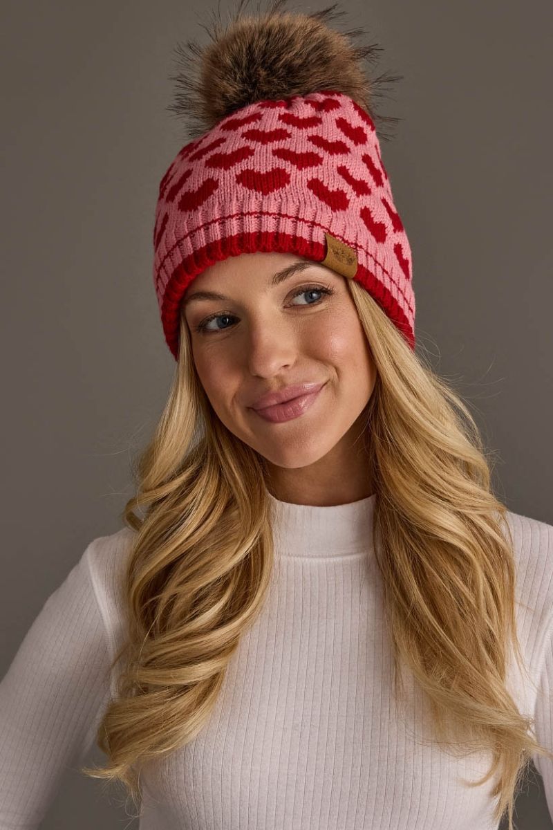 A woman wearing a pink and red knit beanie with a red heart pattern, a brown faux fur pom-pom, and a small brown leather brand tab on the side. The background is a solid neutral gray.