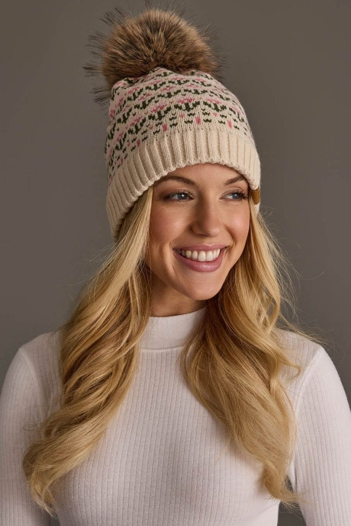 A smiling blonde woman wearing a cream knit beanie with a brown faux-fur pom-pom and a pink and green fair isle pattern, paired with a white ribbed mock-neck top against a solid gray studio background.