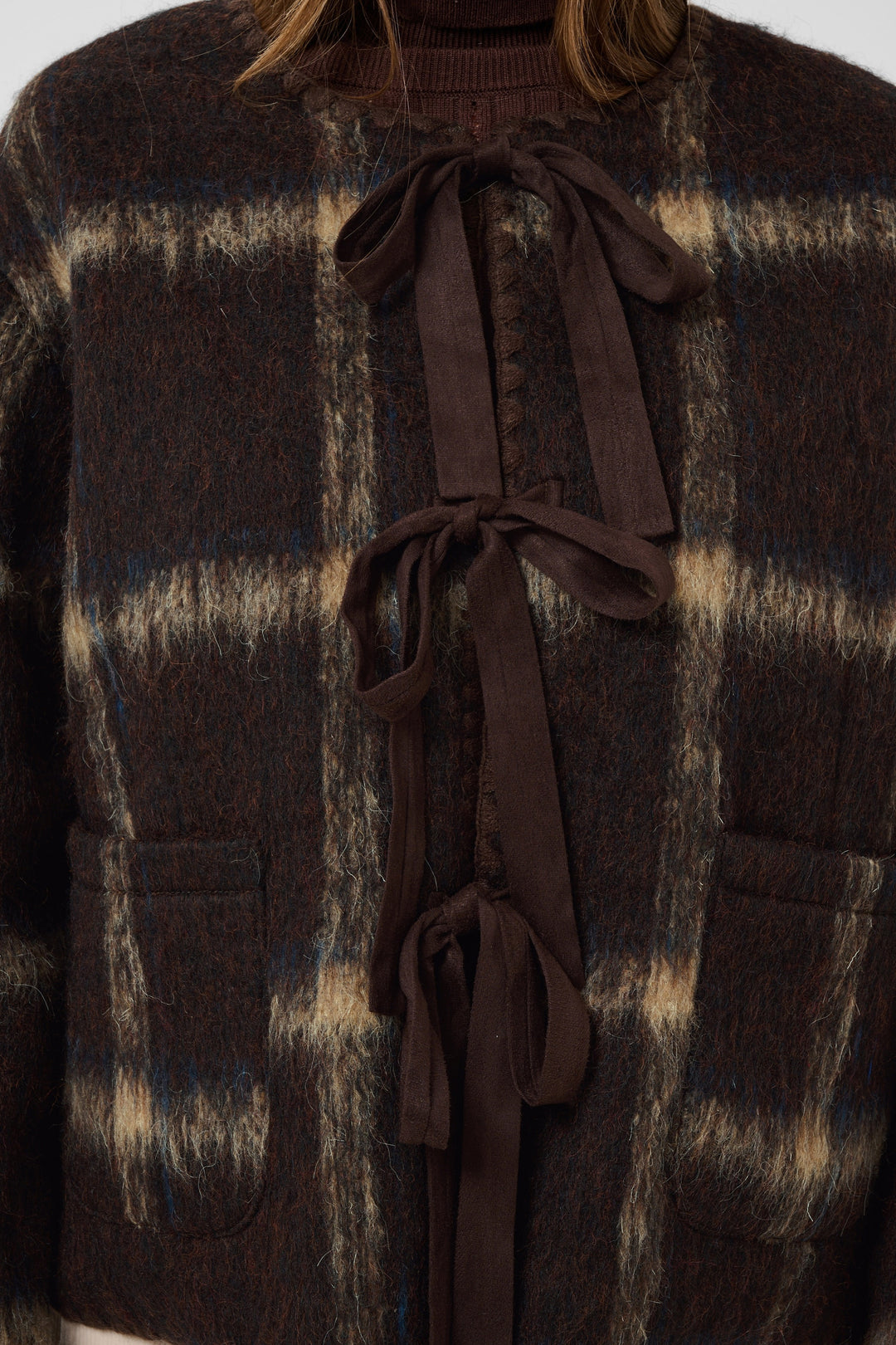 Close-up of a dark brown plaid knit cardigan with a matching dark brown tie-front bow and front patch pockets.