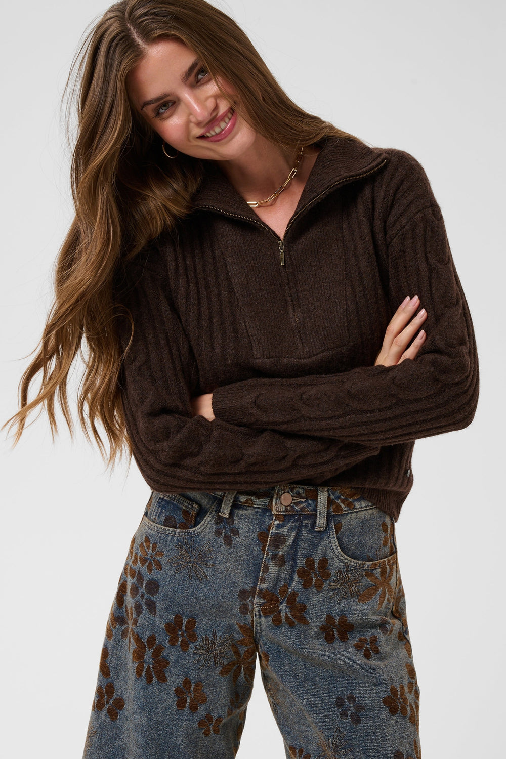A woman modeling a dark brown ribbed quarter-zip mock-neck sweater paired with blue patterned jeans, set against a plain light gray studio background.