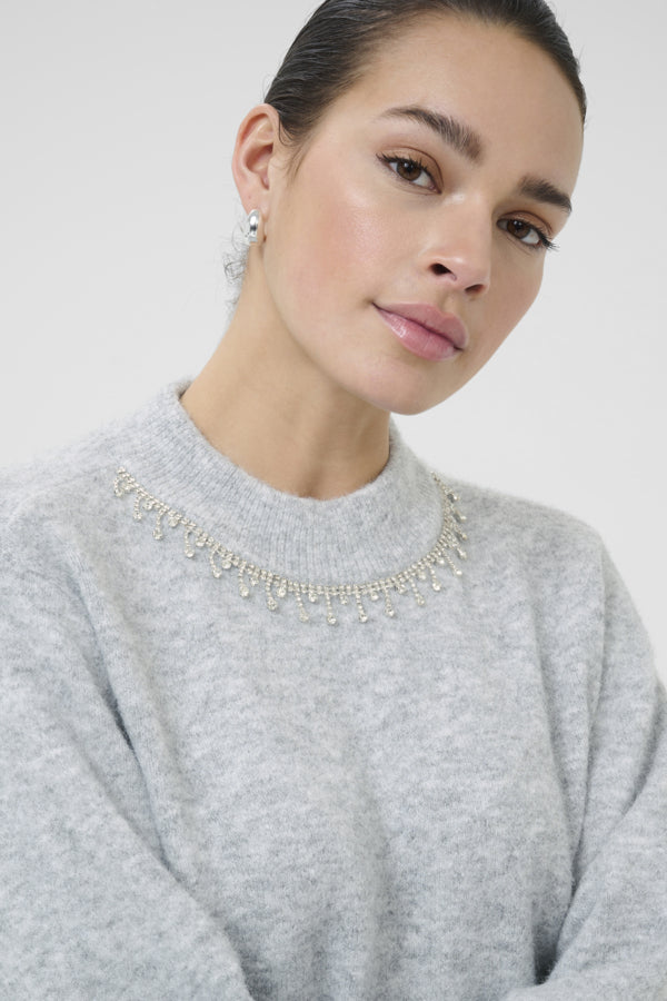 Woman wearing a gray sweater with a decorative necklace against a plain background