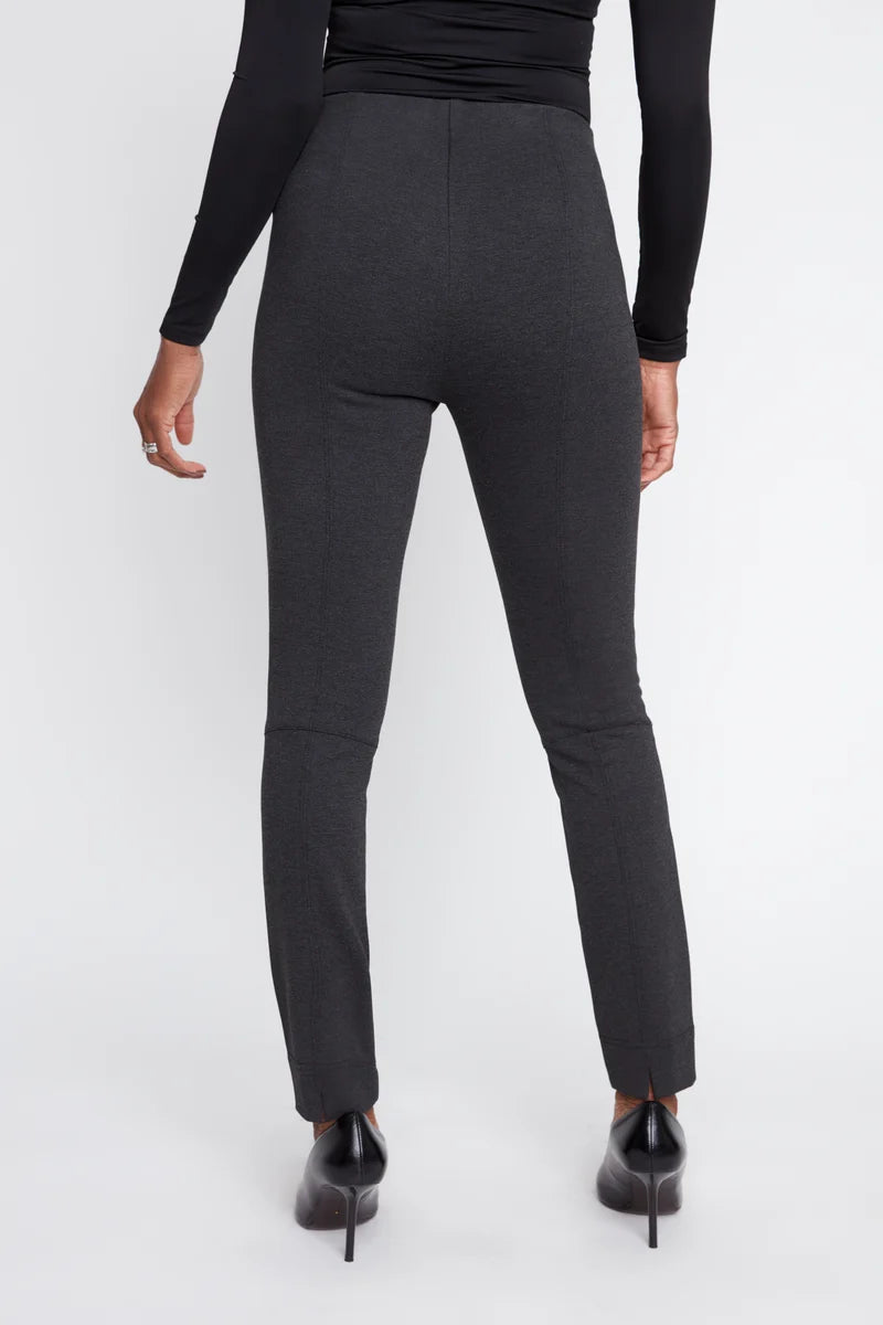 A model shown from the back wearing dark charcoal gray slim-fit trousers, a black long-sleeve top, and black pointed-toe heels against a light gray studio background.